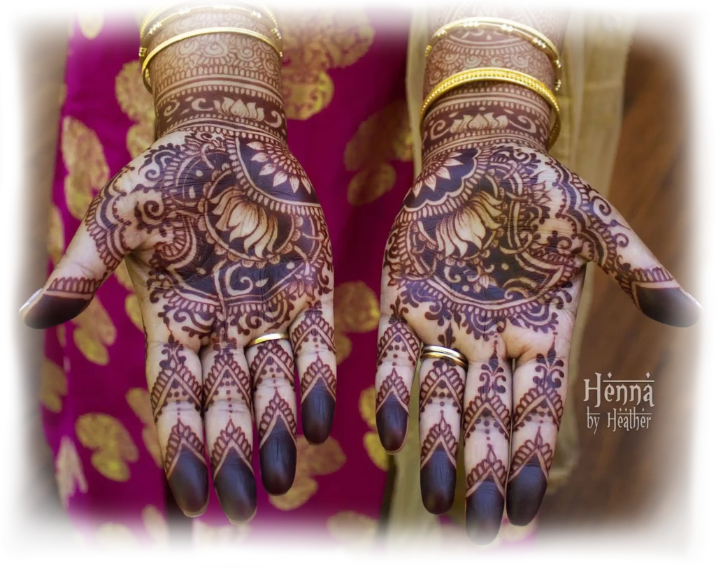 Two palms fully decorated with bridal henna / mehndi, featuring a reverse negative paisley and lotus motif. The fingertips are covered in deep red solid henna with V shape bottom capped fingers. The style is Indian, with traditional motifs and a modern design sensibility. The bride is wearing gold bangle bracelets and a deep fuschia sari with gold accents.
