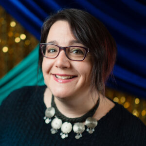 Henna artist Heather Caunt-Nulton, wearing a black necklace with silver medallions, black shirt, glasses, and short hair. In front of the Spring Fling 2026 photo backdrop with glittering gold, royal blue, turquoise, and purple - peacock colors